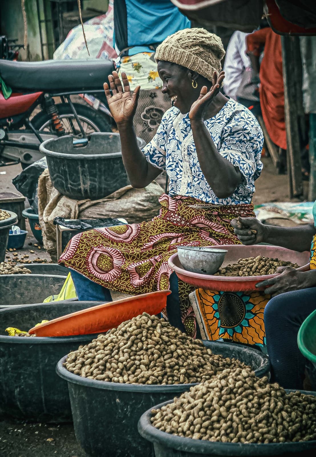 A smiling woman in a head wrap sits behind large bowls of groundnuts at a market.