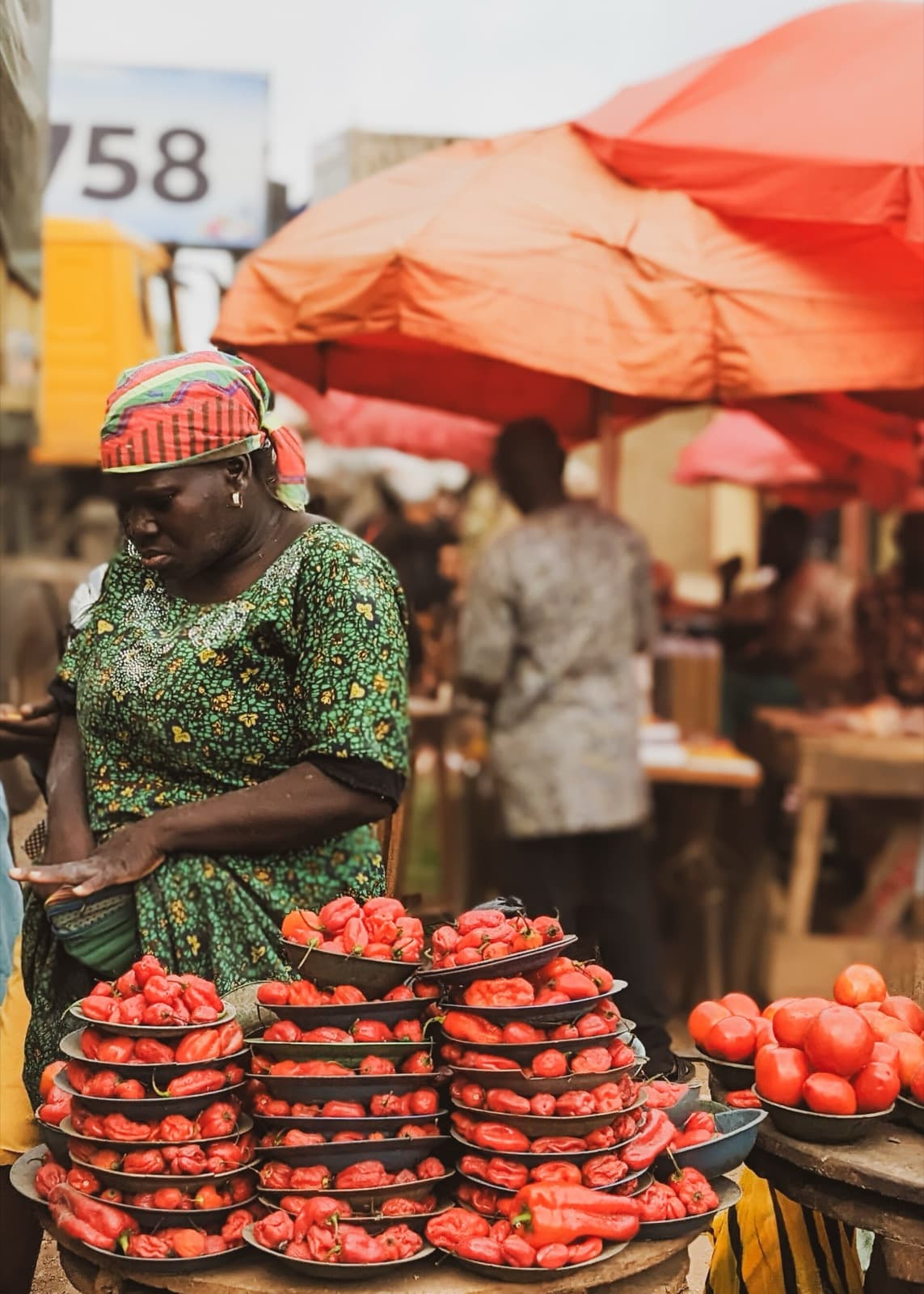 A woman in a green patterned dress arranges trays of red peppers under a red umbrella at Shasha Market.