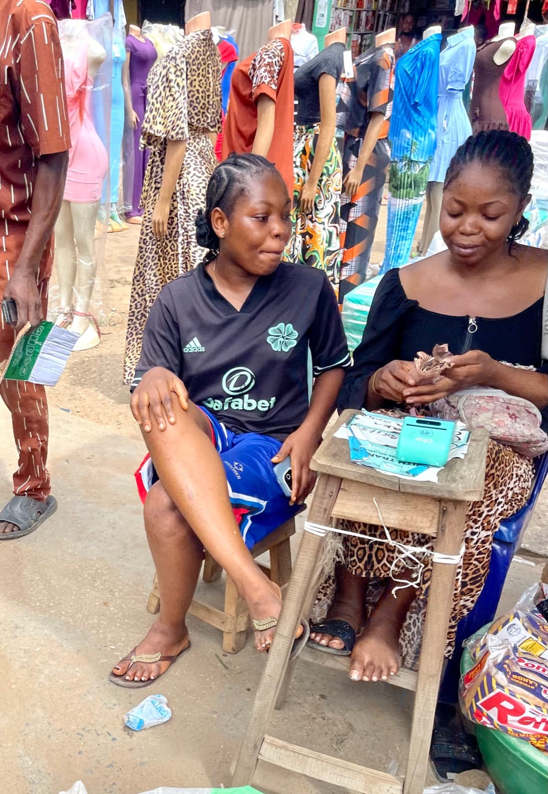 Two women run a POS agent stand at an outdoor market, one counting cash while the other waits.