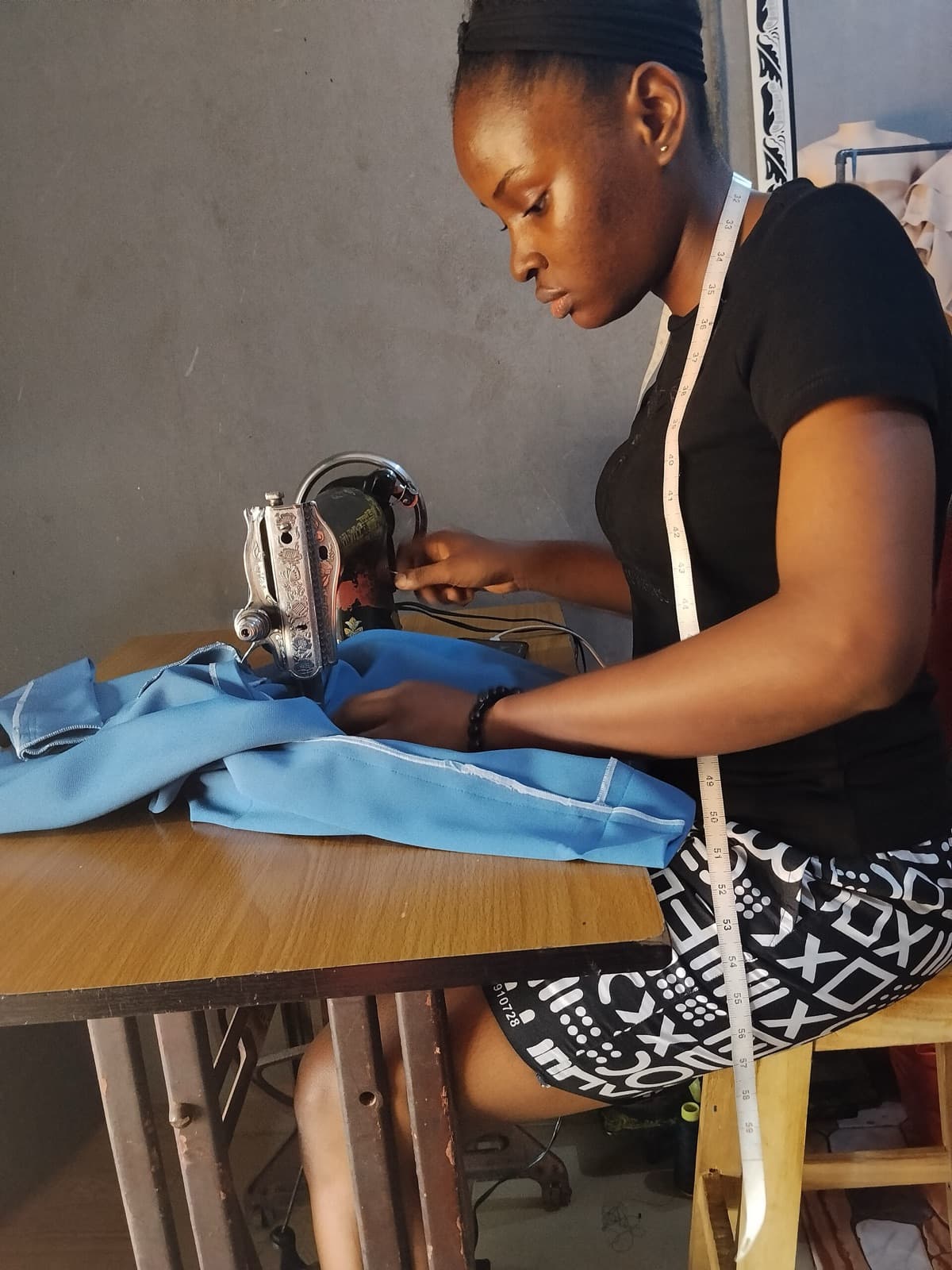 A seamstress with a measuring tape around her neck sews blue fabric at her machine.