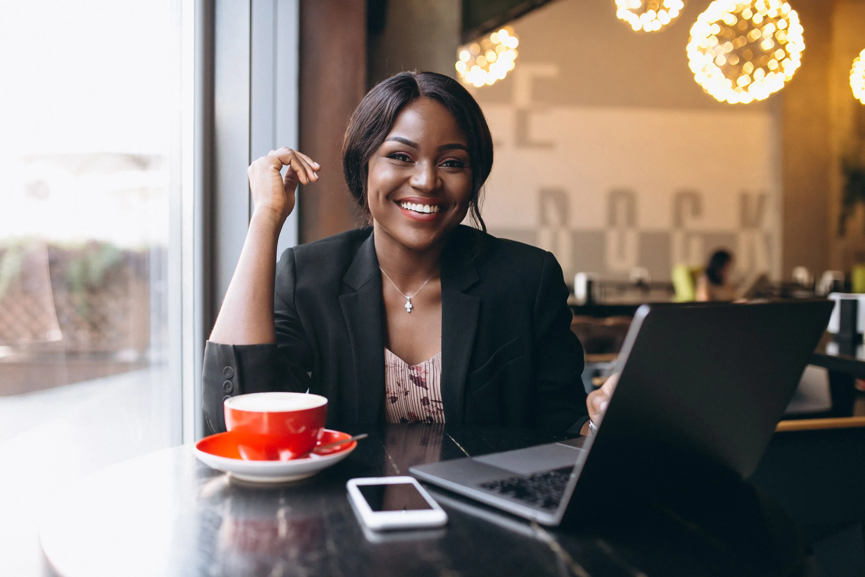 A woman in a black blazer smiles at a café table with a laptop, phone and red cup.