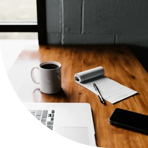 A wooden desk with a mug of coffee, a notepad and pen, a laptop and a phone.