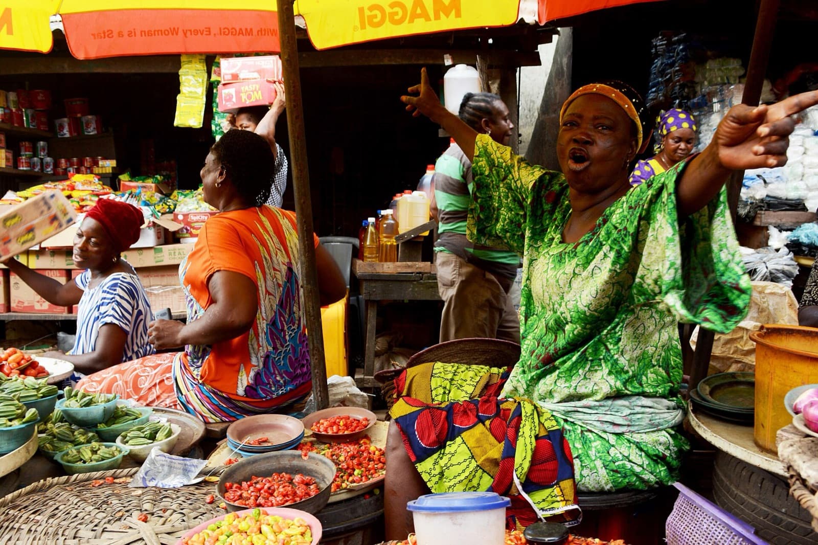 A market trader laughs with her arms raised beside baskets of tomatoes and peppers.