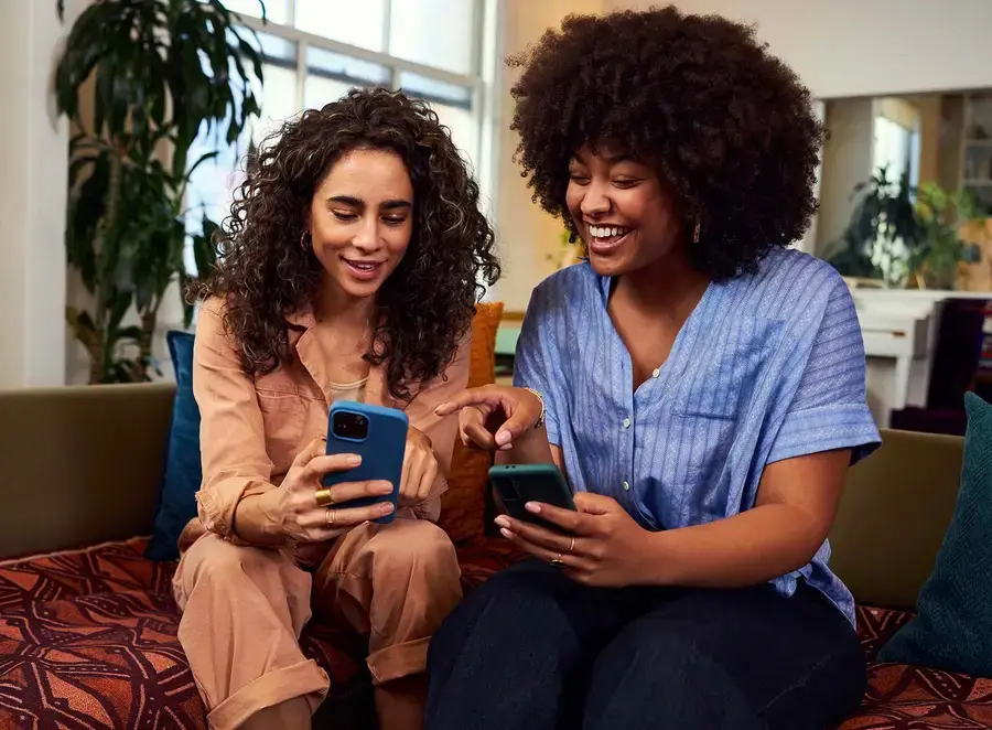 Two friends on a sofa laugh as one points at a referral on the other’s phone.